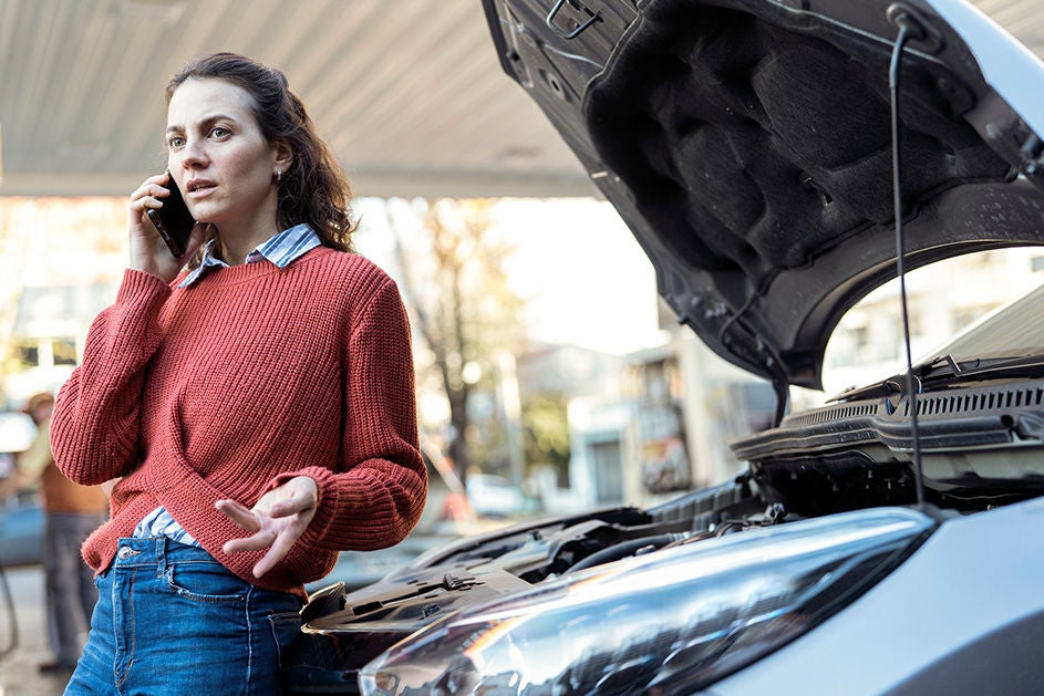 A woman stands beside a broken car, talking on her cell phone with a focused expression.