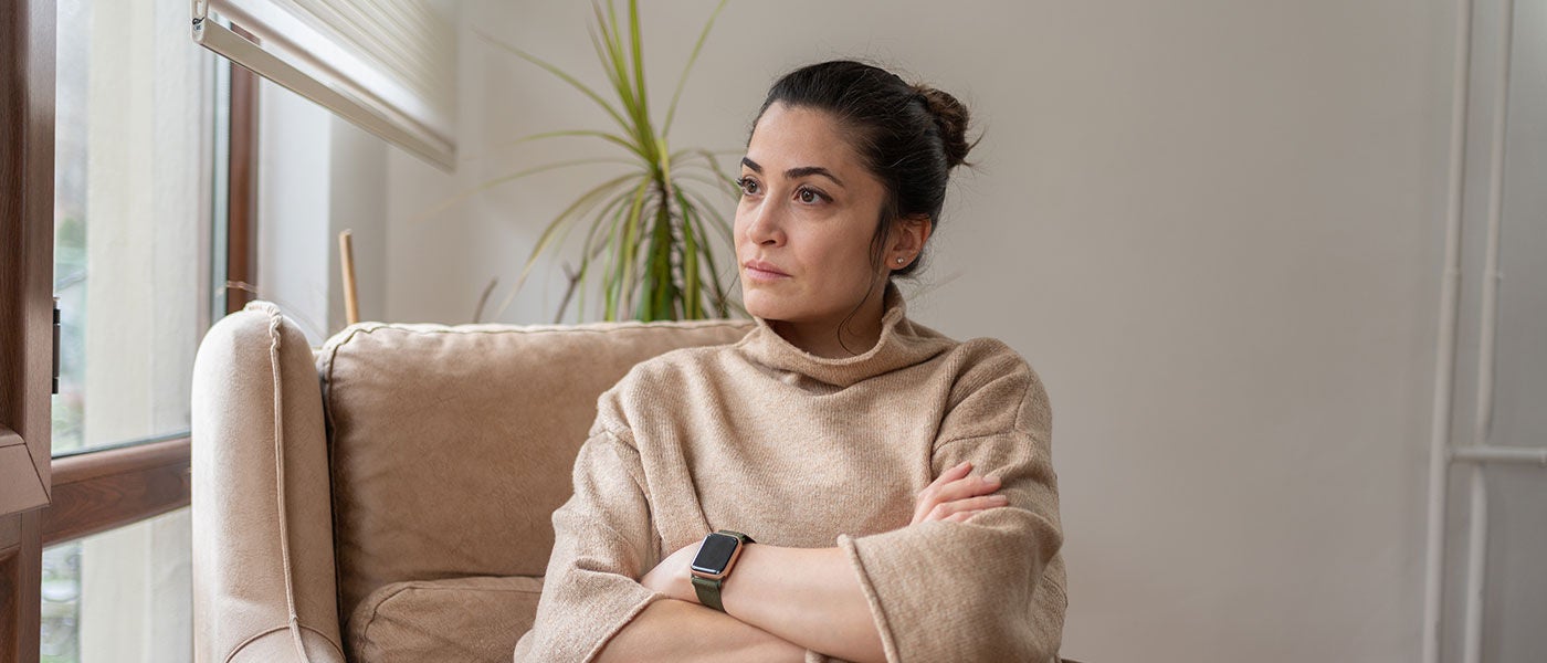Thoughtful woman in a beige sweater sits on a sofa, arms crossed, gazing toward a window in a calm, sunlit room with plants.