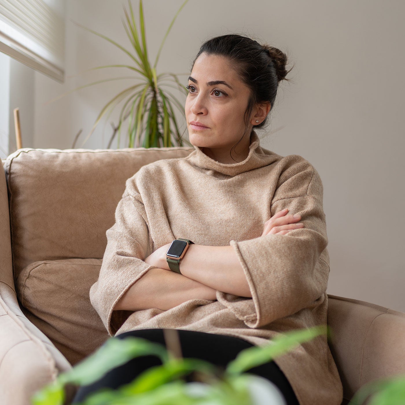 Thoughtful woman in a beige sweater sits on a sofa, arms crossed, gazing toward a window in a calm, sunlit room with plants.