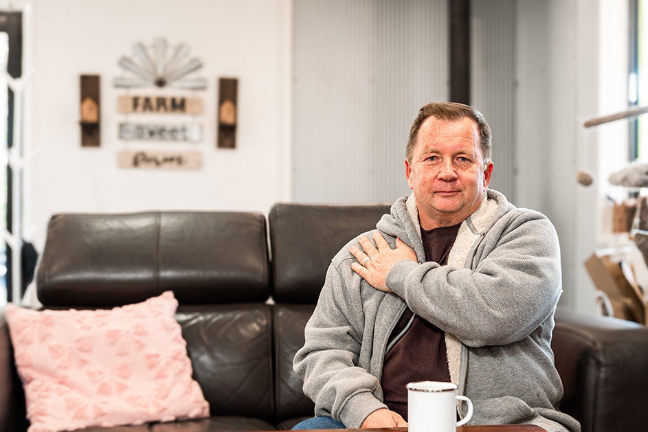 A man sitting on a couch, putting his left hand on his right shoulder.