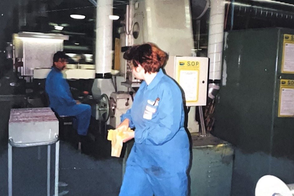 A woman dressed in blue overalls stands beside a machine