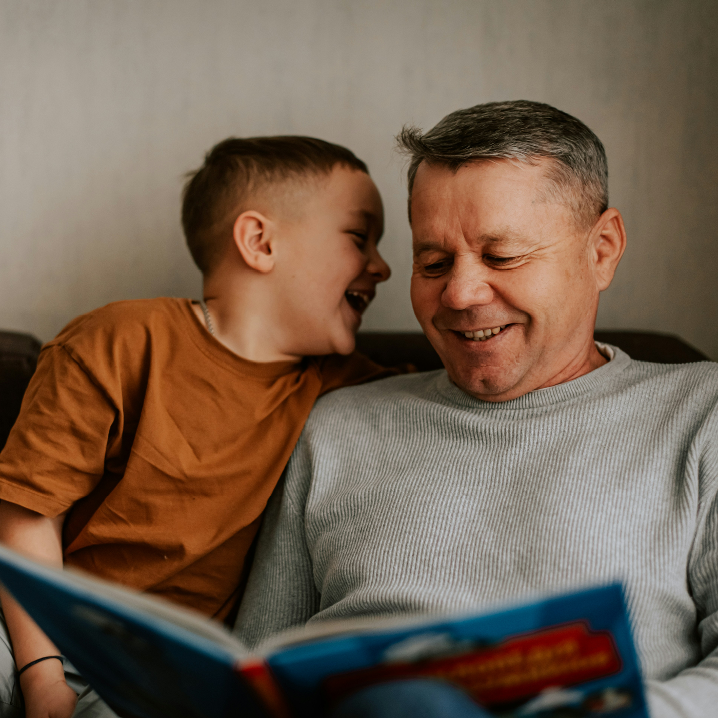 A grandfather and young grandson laughing together on a sofa at home