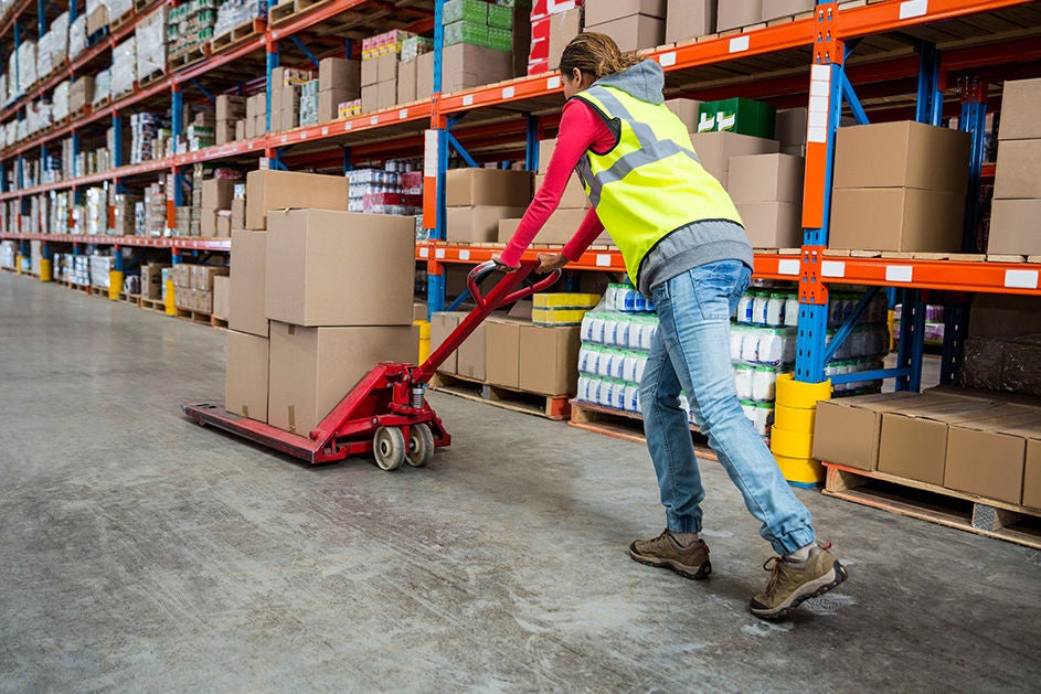 A woman pushing a cart through a spacious warehouse filled with shelves and boxes.
