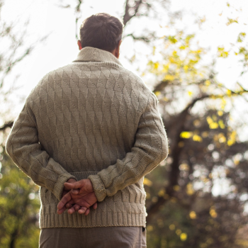 An older man facing away from the camera with his hands crossed behind his back looking out at the a garden