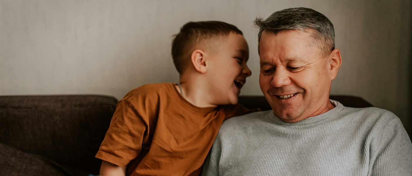 A grandfather and young grandson laughing together on a sofa at home