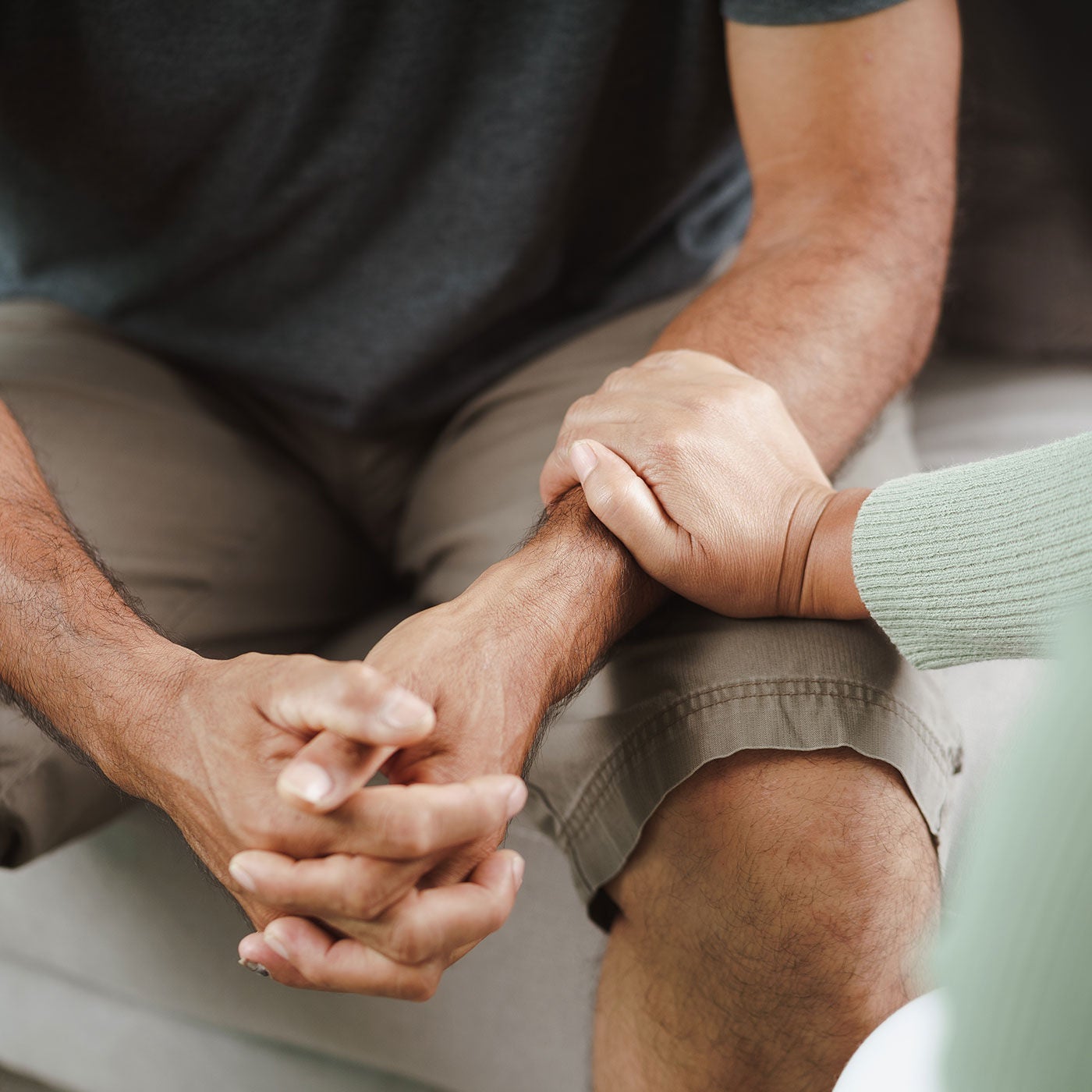 A person sits on a couch with hands clasped while another person rests a hand on their arm in a comforting gesture.