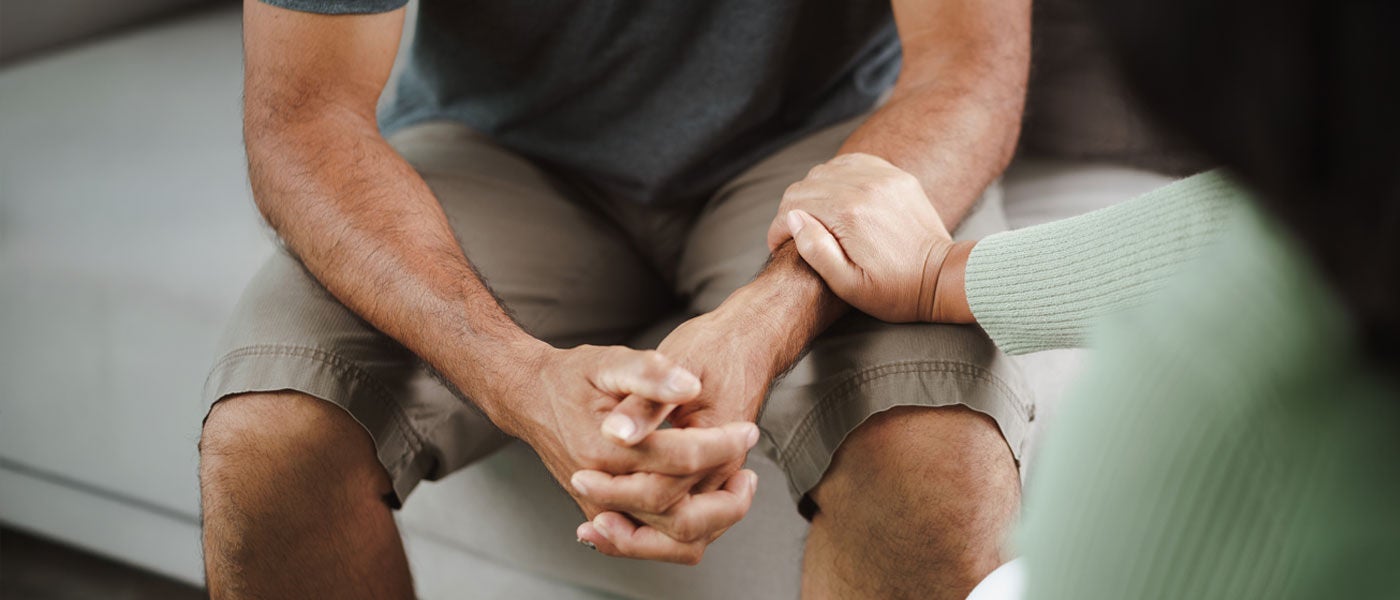 A person sits on a couch with hands clasped while another person rests a hand on their arm in a comforting gesture.