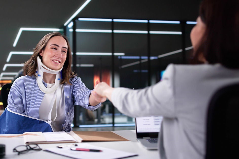 A woman wearing a neck brace shakes hands with another woman, both smiling in a supportive interaction.