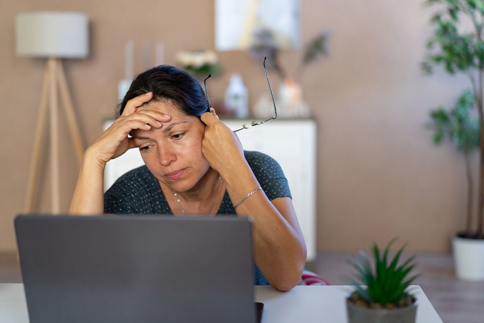 A woman sits at a desk looking at a laptop screen, holding her glasses and resting her head on her hand, appearing stressed or tired.