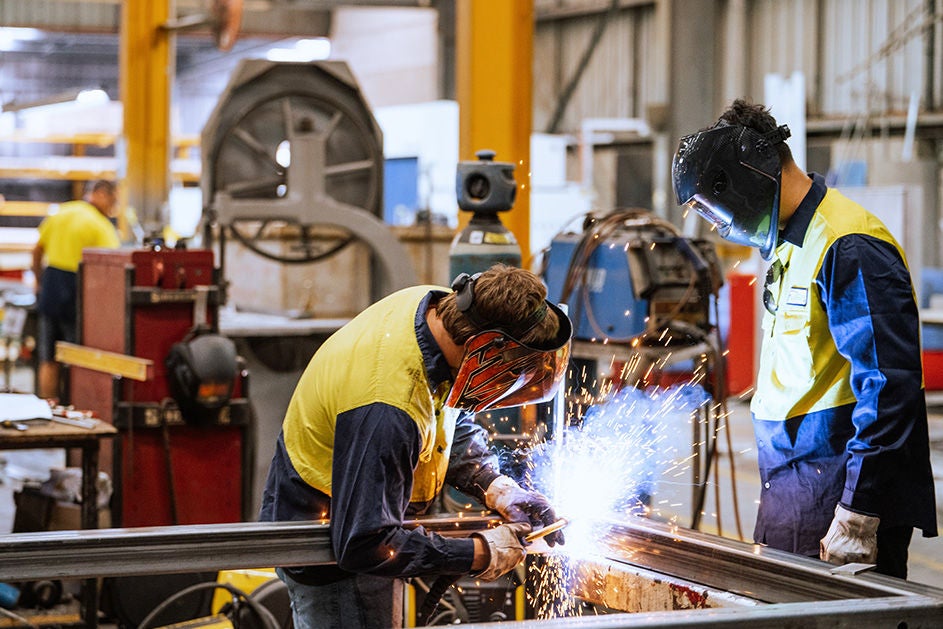 Two men working together on metal fabrication in a factory setting, focused on their tasks with machinery in the background.