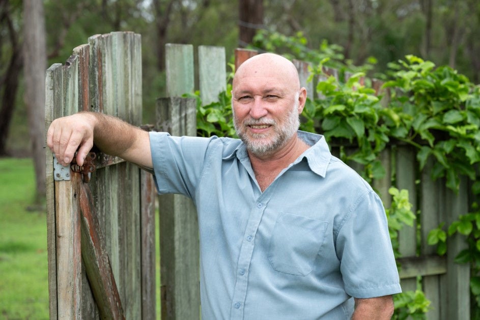 A man casually leans on a fence while holding a garden hose, with plants and flowers in the background.