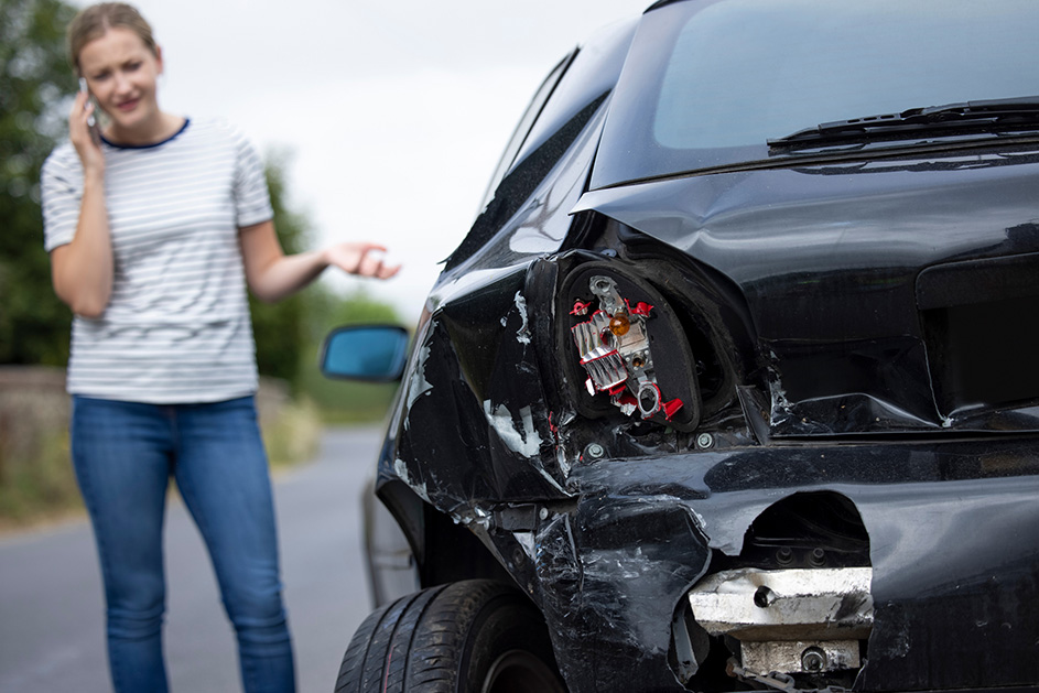 A worried woman speaks on her phone beside a black car with a shattered taillight and crumpled rear bumper after a road accident.