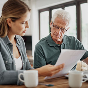 A woman and an elderly man sit at a table, reviewing documents together. The man wears glasses and uses a laptop. The mood is focused and collaborative.