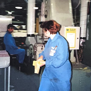 A woman dressed in blue overalls stands beside a machine