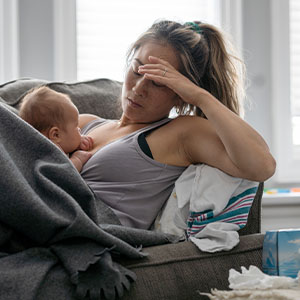 A exhausted new mother resting on a sofa with her newborn baby, with tissues nearby, conveying the physical and emotional toll of the postpartum period