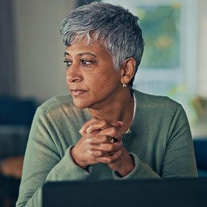 Thoughtful older woman with short gray hair sits behind a laptop, hands clasped, gazing aside in soft natural light at home.