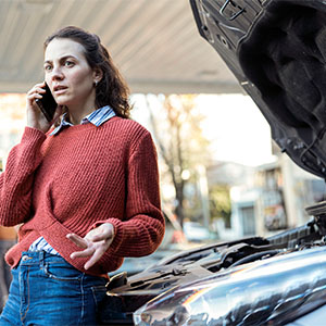 A woman stands beside a broken car, talking on her cell phone with a focused expression.