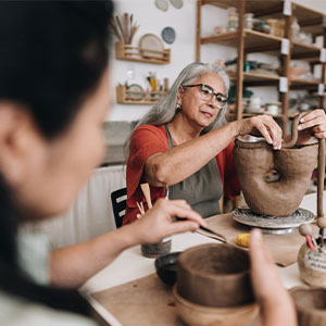 An experienced pottery teacher guides a student in shaping a large clay vessel at a busy ceramics studio.