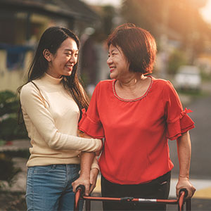 A young woman in a cream sweater supports an older woman in a red blouse using a walker. They smile warmly at each other, bathed in sunset light.