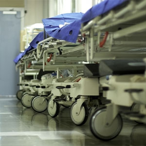 Several empty hospital stretchers with blue sheets are lined up in a hallway near closed double doors.