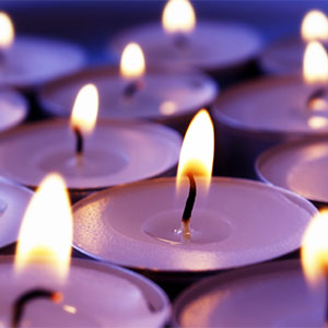 Rows of small tea light candles glow with warm flames against a soft purple-blue background, creating a calm, reflective and peaceful atmosphere.