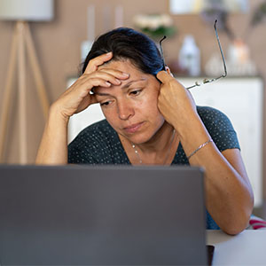 A woman sitting by the desk looking at the laptop screen A woman sits at a desk looking at a laptop screen, holding her glasses and resting her head on her hand, appearing stressed or tired.