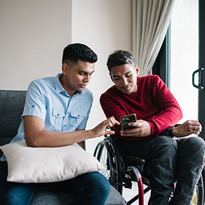 Two friends sit indoors, one using a wheelchair, as they share a smartphone; the relaxed scene conveys friendship, support, and connection.