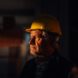 An older male tradesman wearing a hard hat, with a serious expression, in an industrial workplace setting.