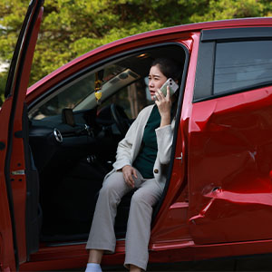 A distressed woman sitting in the open door of a damaged red car, making a phone call after a road accident.