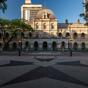 Historic neoclassical building with a domed roof and arched façade glows in warm evening light, framed by trees and a geometric plaza.