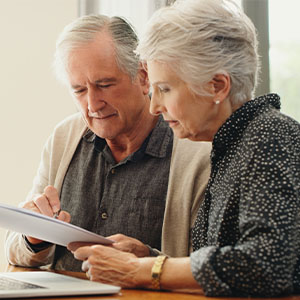 Elderly couple seated together, using a laptop to review paperwork, with focused expressions on their faces.