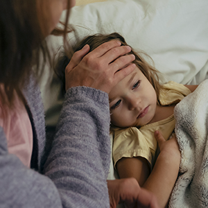 A small child lies in a bed, a woman leans over her placing her hand on her forehead