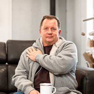 A man sitting on a couch, putting his left hand on his right shoulder.