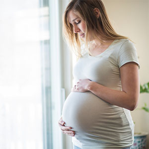 A pregnant woman stands indoors by a window, wearing a light-colored t-shirt and holding her belly with both hands.