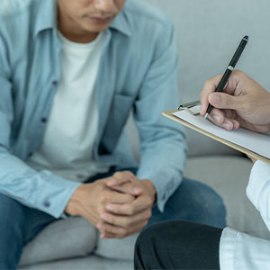 A therapist takes notes on a clipboard as a seated man leans forward with clasped hands, suggesting a calm, focused counseling session.