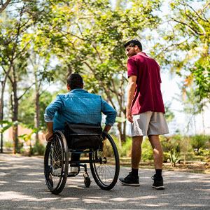Two men enjoying a walk in a sunlit park; one is in a wheelchair, the other walking beside him, surrounded by green trees and pathways.