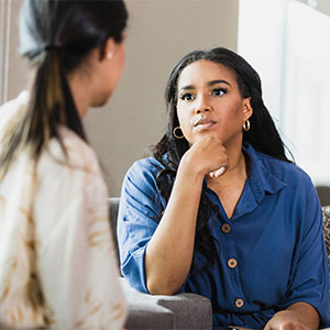 Two women sit indoors having a serious conversation, with one woman facing the camera and listening attentively to the other.