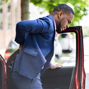 A man in a business suit wincing in back pain as he exits a red car, suggesting injury following a road accident