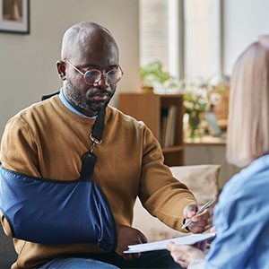 A man with a broken arm sits on a couch, talking to a medical professional.