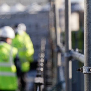 Close-up of a metal scaffold tube secured by a clamp, with a hard-hat worker in a neon safety vest blurred at a construction site.