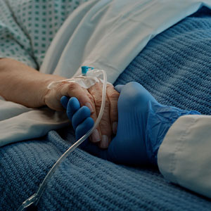 A healthcare worker wearing blue gloves holds the hand of a patient lying in a hospital bed with an IV line attached.