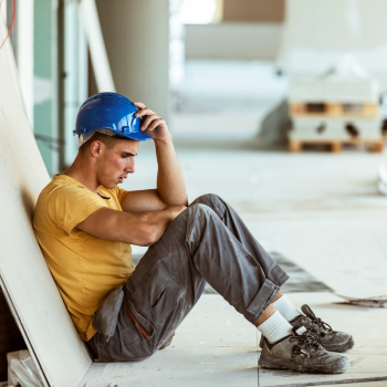A construction worker wearing a hardhat sitting on the floor leaning against a wall wearing 